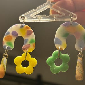 May include: A pair of colorful, handmade earrings featuring a rainbow-colored, speckled, half-circle shape with a yellow flower charm and a swirl-patterned dangle. The other earring features a green flower charm and a swirl-patterned dangle.