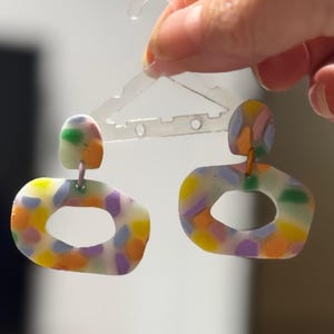 May include: A pair of colorful, abstract, handmade earrings with a white base and a confetti-like pattern of purple, orange, yellow, and green. The earrings are shaped like an oval with a hole in the center.
