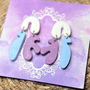 May include: A pair of handmade earrings featuring three different shapes: a curved shape, a squiggly shape, and a long, thin shape. The earrings are made of polymer clay and are painted in shades of light blue, purple, and white.