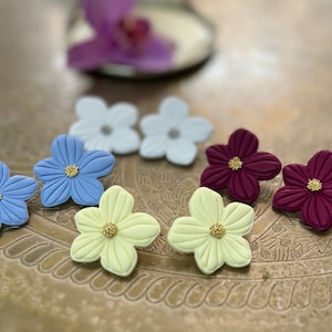 May include: Six pairs of polymer clay flower earrings in various colors, including light blue, yellow, white, and burgundy. Each flower has a gold center.