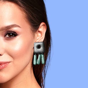 May include: A pair of earrings with a square-shaped pendant and dangling teardrop-shaped beads. The pendant is a dark gray color with a black circle in the center. The beads are a light teal color.