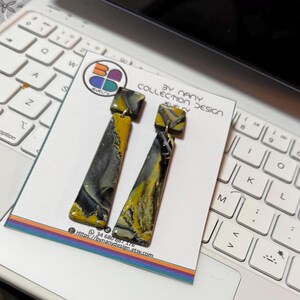 May include: A pair of long, black, gray, and yellow dangle earrings with a square top. The earrings are on a white card with the text "BY NANY COLLECTION DESIGN JEWELY" and a website address.