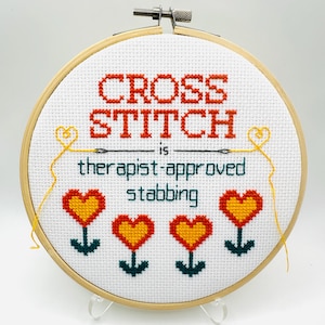 May include: A cross-stitch embroidery hoop with the text "CROSS STITCH is therapist-approved stabbing." The design includes orange text, green text, and yellow heart-shaped flowers. The hoop is made of wood and has a metal tightening mechanism.