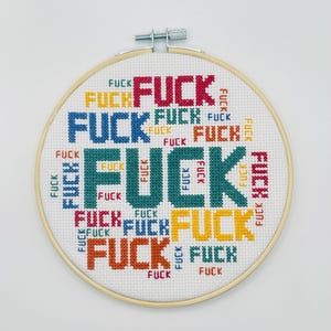 Puede incluir: Un bastidor de bordado a punto de cruz con la palabra "FUCK" repetida en varios colores y tamaños. La palabra está bordada en rojo, azul, verde, naranja y amarillo sobre un fondo blanco. El bastidor de madera tiene un cierre metálico.