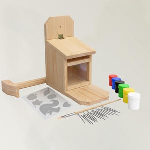 Könnte beinhalten: Ein Eichhörnchenfutter-Set aus Holz mit natürlicher Holzoberfläche. Das Set enthält einen Futterautomaten, Farbtöpfe in verschiedenen Farben, einen Pinsel, Nägel und Schablonen mit Eichhörnchen- und Blattmotiven. Der Futterautomat hat ein Klappdach und ein klares Sichtfenster.