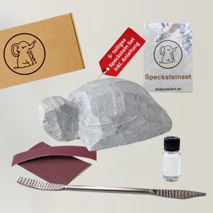 Könnte beinhalten: Ein 5-teiliges Speckstein-Set bestehend aus einem rohen grauen Specksteinblock, einer Metallraspel, zwei dunkelroten Schleifpapierstücken und einer kleinen klaren Ölflasche, inklusive braunem Karton und einer Karte mit Elefanten-Logo.