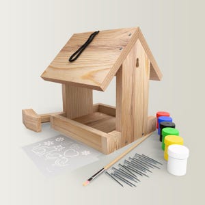 May include: A wooden bird feeder kit featuring a natural grain finish, a black hanging cord, a stencil sheet with various shapes, a wooden paintbrush, a set of small metal nails, and several small paint pots in red, blue, black, green, yellow, and white.