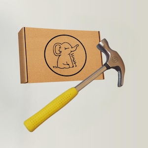 Op de afbeelding: Een klauwhamer met een gele rubberen grip en een zilveren kop. Een bruine kartonnen doos met een zwart cirkelvormig logo van een olifant staat op de achtergrond. Het logo bevat ook de tekst "He I".