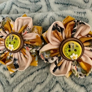 Pode incluir: Duas presilhas de cabelo de flores de tecido feitas à mão. Cada flor apresenta pétalas em amarelo, rosa e padrões florais, com um botão de madeira no centro. O botão é verde com uma pequena flor branca e um detalhe de abelha.
