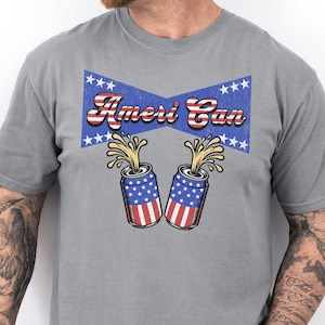 Puede incluir: Una camiseta gris con un gráfico de estilo vintage de dos latas de cerveza con la bandera estadounidense y el texto "Ameri Can" encima.