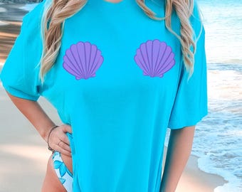 May include: A turquoise t-shirt with two purple seashell designs. The shirt is a solid colour and has a relaxed fit. The image is taken on a beach with the sea in the background.