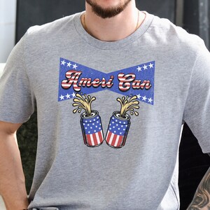 Puede incluir: Una camiseta gris con un gráfico de estilo vintage de dos latas con temática de la bandera estadounidense con el texto "Ameri Can" encima.