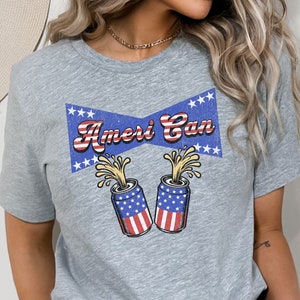 Puede incluir: Camiseta gris con un gráfico azul y rojo de dos latas con temática de la bandera estadounidense con el texto "Ameri Can" en una fuente retro.