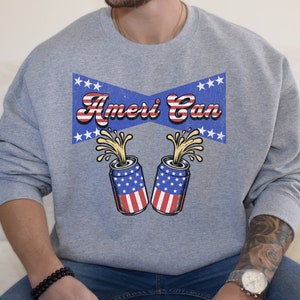 Puede incluir: Una sudadera gris con un gráfico de estilo vintage de dos latas de cerveza con temática de la bandera estadounidense con el texto "Ameri Can" encima.