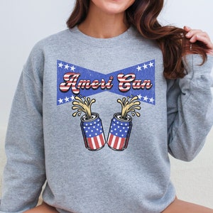 May include: A gray sweatshirt featuring a graphic design with the text "Ameri Can" in red and white, set against a blue banner with white stars. Two cans with American flag designs are depicted, with yellow liquid spilling out.