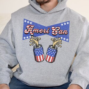 Puede incluir: Una sudadera gris con un diseño de cinta azul y blanca que dice "Ameri Can" con dos latas rojas, blancas y azules con un líquido amarillo que sale a borbotones.