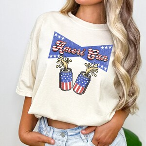 Puede incluir: Una camiseta blanca con un gráfico de estilo vintage de dos latas con temática de la bandera estadounidense con el texto "Ameri Can" en una cinta azul y roja.
