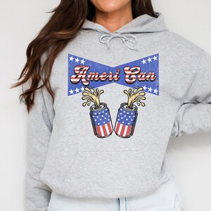 Puede incluir: Una sudadera gris con un diseño azul y rojo con dos latas de cerveza con banderas americanas y el texto "Ameri Can".