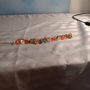 May include: A bracelet featuring a thin gold-toned metal chain adorned with a series of small, round buttons in shades of orange, white, and green. The buttons are attached at intervals along the chain, creating a layered and textured appearance.