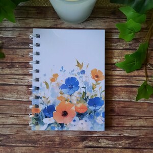 Könnte beinhalten: Ein weißes Spiralnotizbuch mit einem Aquarell-Blumenmuster. Das Design zeigt blaue und orangefarbene Blumen mit grünen Stielen und Blättern.