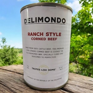 May include: A white can of Delimondo Ranch Style Corned Beef. The can has a red and black label with the brand name and product description. The can is sitting on a wooden surface. The text on the can reads: "Made from 100% cattle beef. This premium, long-strand corned beef is cooked the old-fashioned way. Specially cured and seasoned to perfection. Tastes Like Home®"