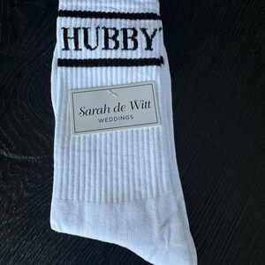May include: White athletic socks with black stripes and the word "HUBBY" in black block letters. A small tag reads "Sarah de Witt WEDDINGS". The socks are ribbed and appear to be new.