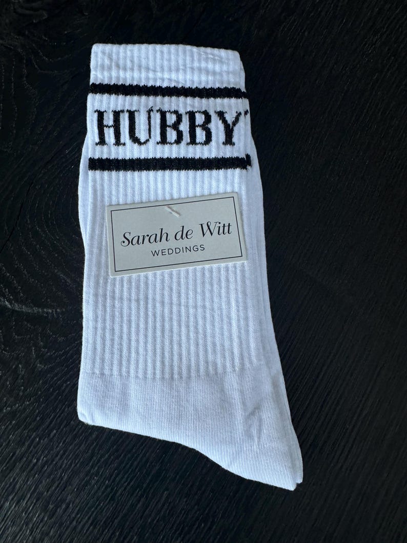 May include: White athletic socks with black stripes and the word "HUBBY" in black block letters. A small tag reads "Sarah de Witt WEDDINGS". The socks are ribbed and appear to be new.