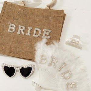 May include: A collection of bridal accessories, including a brown burlap tote bag with the word "BRIDE" in pearl letters, heart-shaped sunglasses with pearl accents, a white feather fan with pearl lettering, and a hair clip.