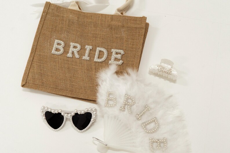May include: A collection of bridal accessories, including a brown burlap tote bag with the word "BRIDE" in pearl letters, heart-shaped sunglasses with pearl accents, a white feather fan with pearl lettering, and a hair clip.