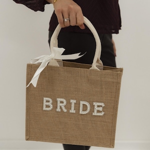 May include: A tan burlap tote bag with the word "BRIDE" spelled out in pearl-like letters. The bag has cream-colored handles and a white ribbon tied in a bow. The bag is being held by a person.