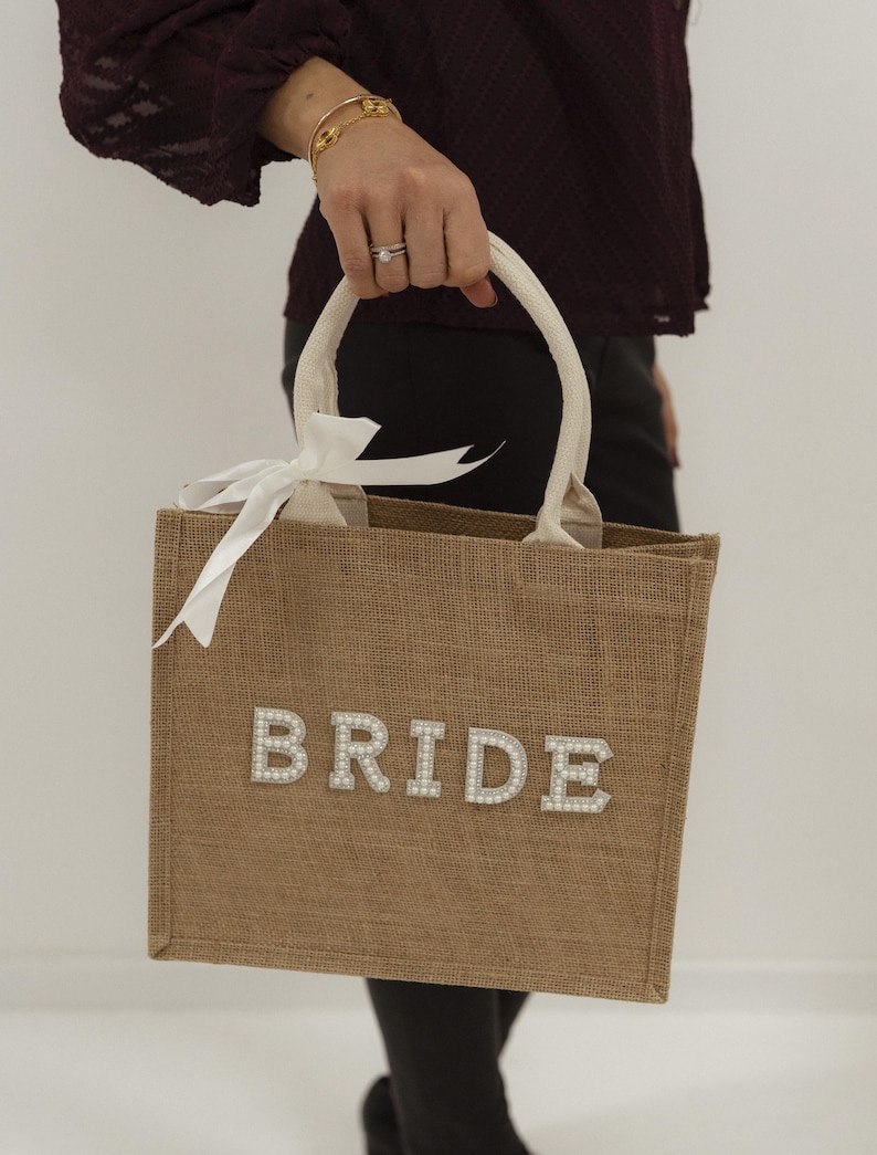 May include: A tan burlap tote bag with the word "BRIDE" spelled out in pearl-like letters. The bag has cream-colored handles and a white ribbon tied in a bow. The bag is being held by a person.
