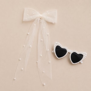 May include: A white tulle bow with pearl accents and long, flowing tails, paired with heart-shaped sunglasses adorned with pearls and black lenses. The accessories are set against a neutral background.