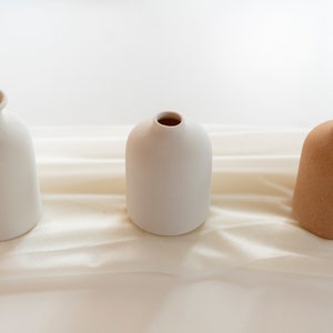 May include: Three ceramic vases, two white and one brown, arranged in a row on a white fabric background.