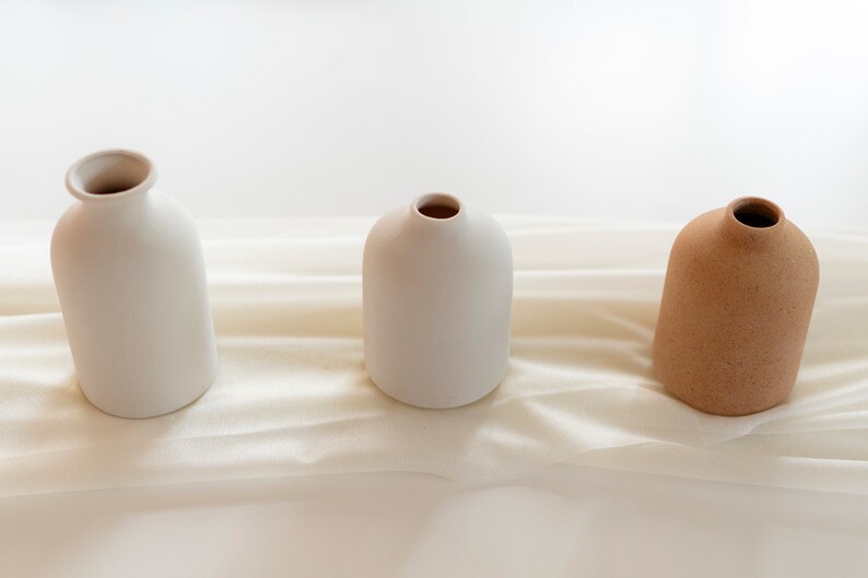 May include: Three ceramic vases, two white and one brown, arranged in a row on a white fabric background.