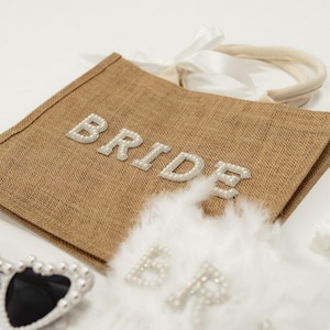 May include: A brown burlap tote bag with the word "BRIDE" spelled out in pearl-like letters. The bag is accompanied by heart-shaped sunglasses with pearl accents, a white feather boa, and a hair clip.