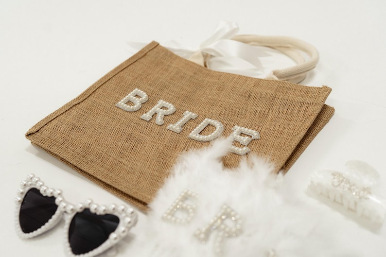 May include: A brown burlap tote bag with the word "BRIDE" spelled out in pearl-like letters. The bag is accompanied by heart-shaped sunglasses with pearl accents, a white feather boa, and a hair clip.