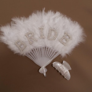 Könnte beinhalten: Ein weißer Federfächer mit dem Wort „BRIDE“ in glitzernden Buchstaben. Eine passende Haarspange ist ebenfalls vorhanden. Der Fächer und die Spange sind wahrscheinlich Brautaccessoires.