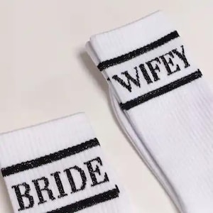K&ouml;nnte beinhalten: Wei&szlig;e Socken mit schwarzem Text, der "BRIDE" und "WIFEY" lautet. Die Socken haben einen schwarzen Streifen oben und unten am Text. Die Socken sind aus einem weichen, gerippten Material.