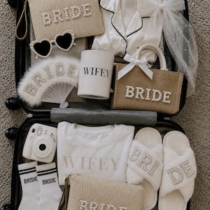 May include: A suitcase filled with bridal accessories. Items include a white satin pajama set, a small tote bag, a fan, and two small pouches, all with the word "BRIDE" spelled out in pearls. Also included are heart-shaped sunglasses, a mug that says "WIFEY", socks, and fluffy slippers.