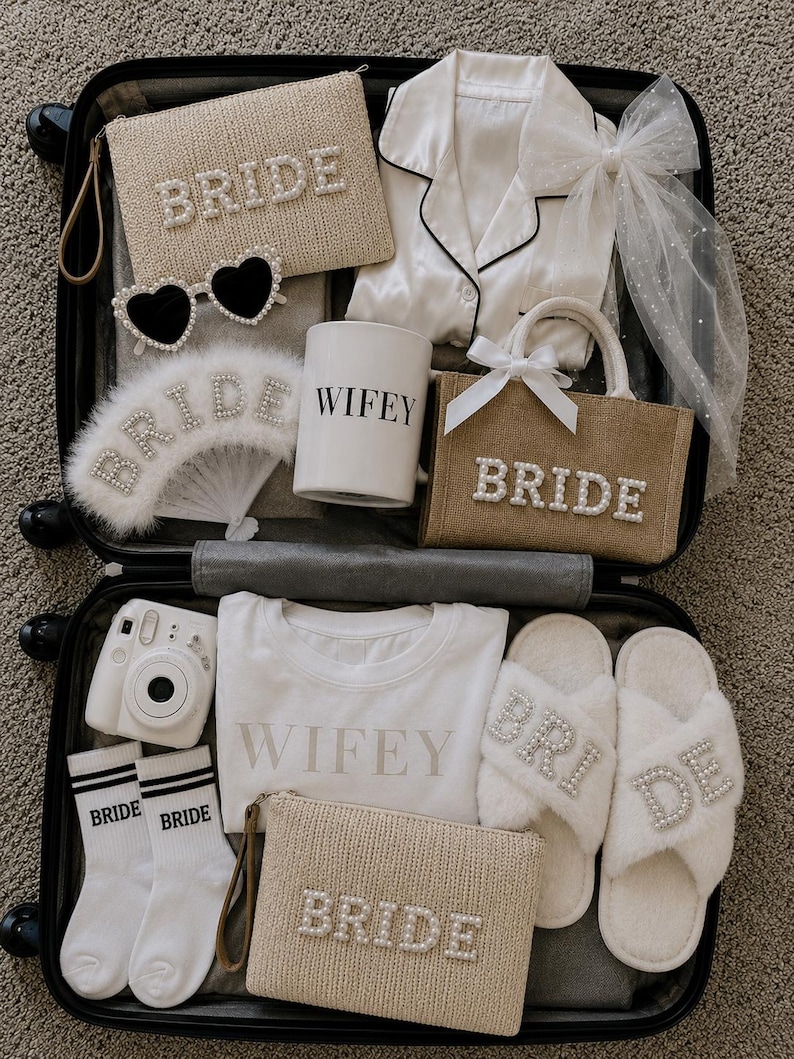 May include: A suitcase filled with bridal accessories. Items include a white satin pajama set, a small tote bag, a fan, and two small pouches, all with the word "BRIDE" spelled out in pearls. Also included are heart-shaped sunglasses, a mug that says "WIFEY", socks, and fluffy slippers.