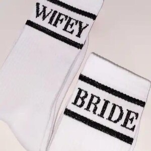 K&ouml;nnte beinhalten: Wei&szlig;e Socken mit schwarzem Text und Streifen. Auf einer Socke steht "WIFEY" und auf der anderen "BRIDE". Die Socken sind wahrscheinlich f&uuml;r eine Hochzeit oder eine Brautveranstaltung gedacht.