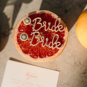 May include: A halved grapefruit with a sparkling "Bride" word charm. The charm is made of small, clear crystals and is attached to two decorative circular accents. A partially visible orange is on the right, and a card is at the bottom.