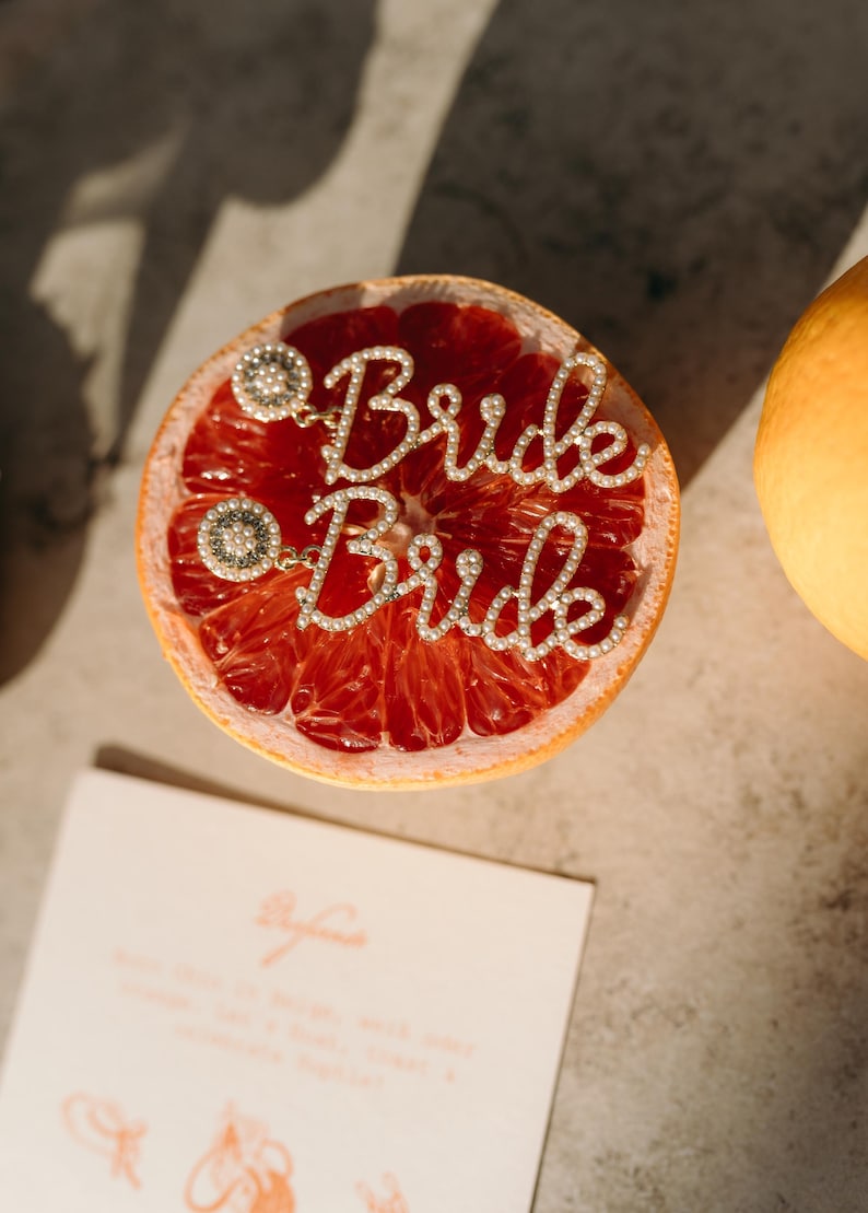 May include: A halved grapefruit with a sparkling "Bride" word charm. The charm is made of small, clear crystals and is attached to two decorative circular accents. A partially visible orange is on the right, and a card is at the bottom.