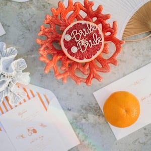 May include: A coral-colored decorative piece with a grapefruit slice, adorned with the word "Bride" in sparkling letters. The image also includes a white fan, invitation cards, and an orange, suggesting a wedding or bridal theme.