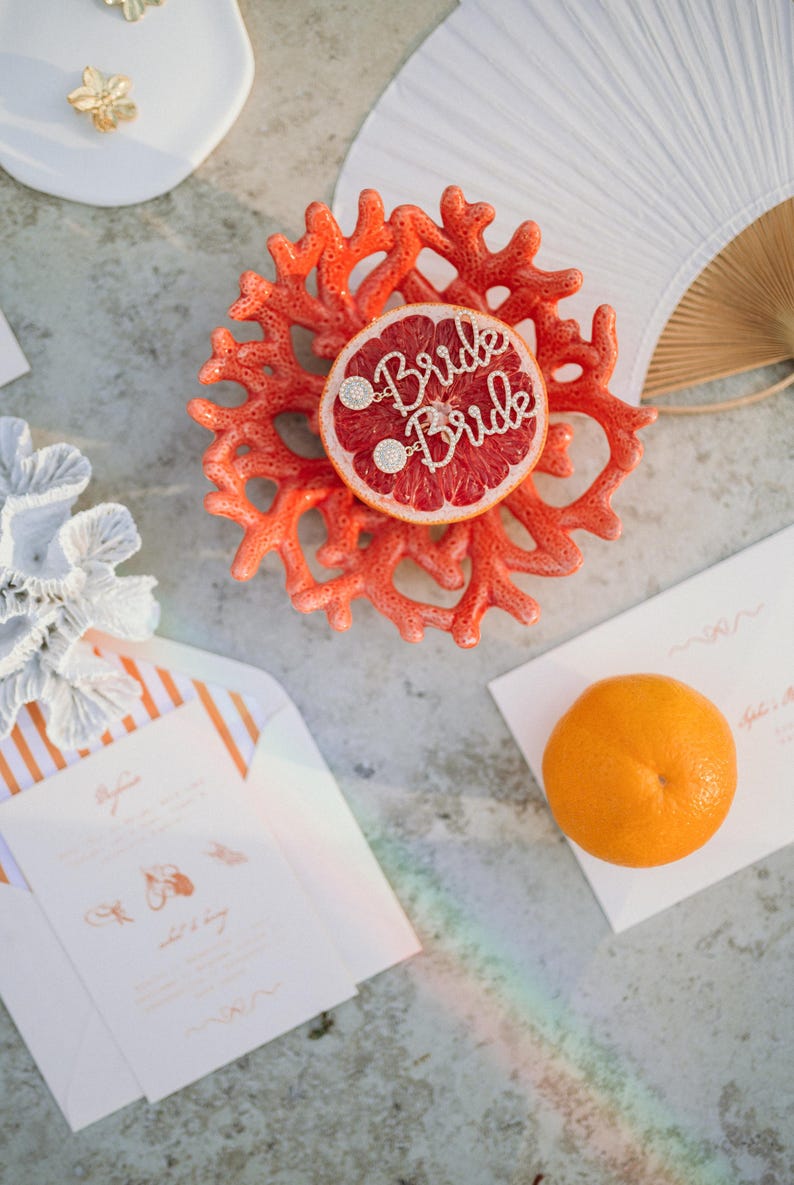 May include: A coral-colored decorative piece with a grapefruit slice, adorned with the word "Bride" in sparkling letters. The image also includes a white fan, invitation cards, and an orange, suggesting a wedding or bridal theme.