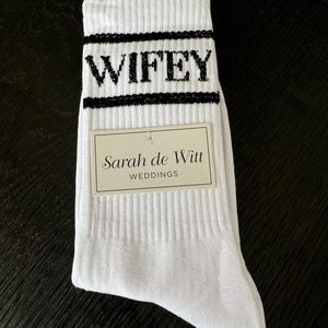 K&ouml;nnte beinhalten: Wei&szlig;e gerippte Socken mit dem Wort "WIFEY" in schwarzen Blockbuchstaben. Die Socken haben einen schwarzen Streifen &uuml;ber und unter dem Wort. Ein rechteckiges Etikett mit dem Text "Sarah de Witt WEDDINGS" ist angebracht.