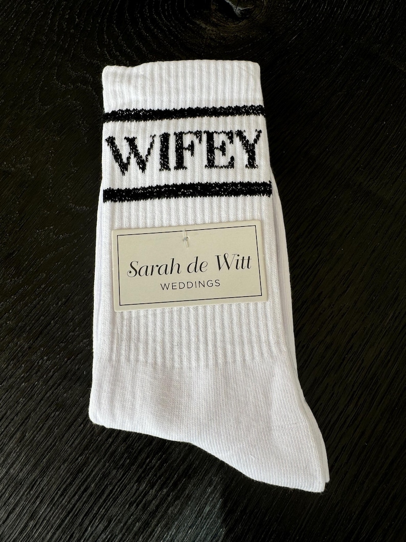 K&ouml;nnte beinhalten: Wei&szlig;e gerippte Socken mit dem Wort "WIFEY" in schwarzen Blockbuchstaben. Die Socken haben einen schwarzen Streifen &uuml;ber und unter dem Wort. Ein rechteckiges Etikett mit dem Text "Sarah de Witt WEDDINGS" ist angebracht.