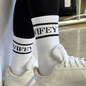 K&ouml;nnte beinhalten: Wei&szlig;e Sportsocken mit dem Wort "WIFEY" in schwarzen Blockbuchstaben. Die Socken werden mit schwarzen Leggings und wei&szlig;en Turnschuhen getragen. Die Turnschuhe haben wei&szlig;e Schn&uuml;rsenkel und das Adidas-Logo.