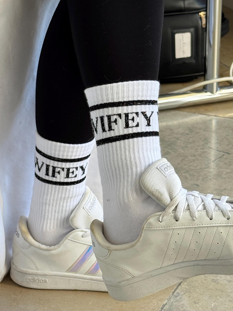 K&ouml;nnte beinhalten: Wei&szlig;e Sportsocken mit dem Wort "WIFEY" in schwarzen Blockbuchstaben. Die Socken werden mit schwarzen Leggings und wei&szlig;en Turnschuhen getragen. Die Turnschuhe haben wei&szlig;e Schn&uuml;rsenkel und das Adidas-Logo.