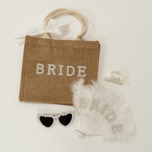 May include: A collection of bridal accessories, including a burlap tote bag with "BRIDE" in rhinestone letters, heart-shaped sunglasses, a feather fan, and a hair clip. The fan and bag also feature the word "BRIDE".